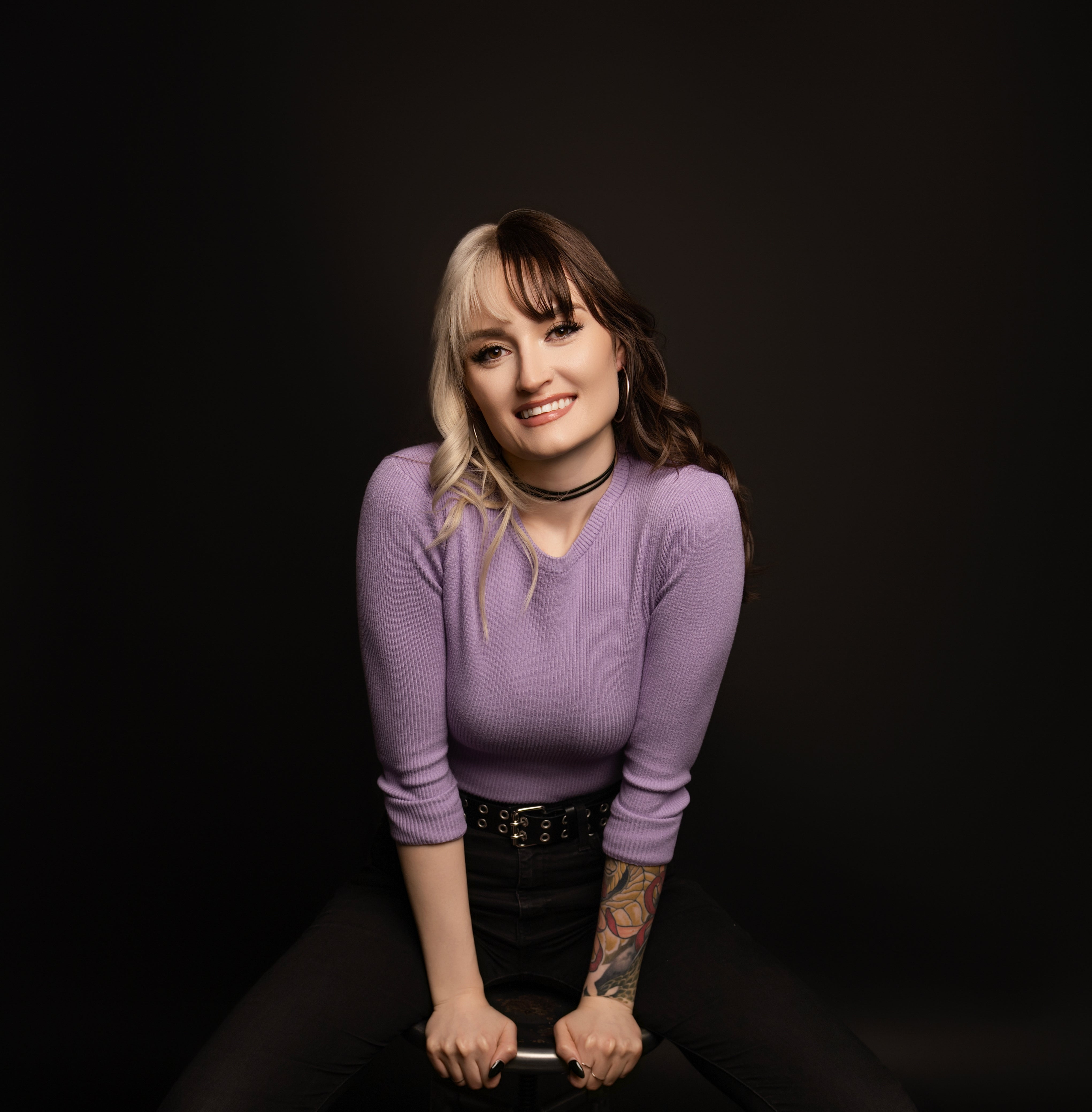 Woman in a purple sweater sitting on a black stool against a dark background
