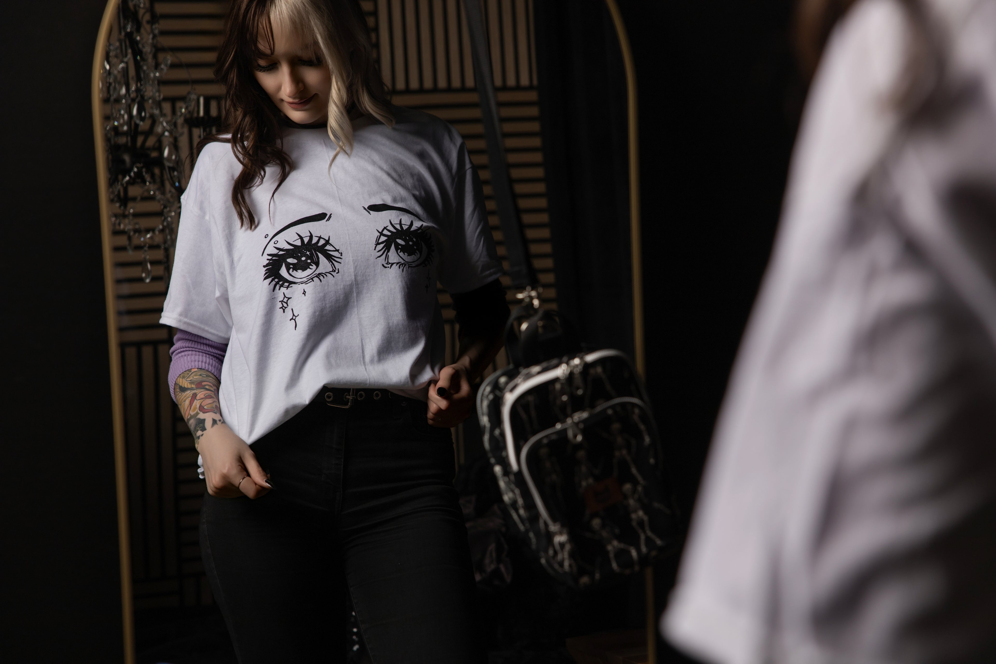Person wearing a white t-shirt with eye designs, a black bag is on a mirror in a dimly lit room.