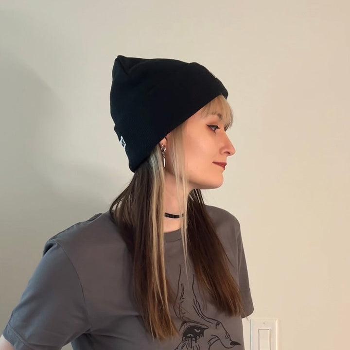 Person wearing a black beanie and gray shirt with a graphic design, standing against a plain wall.